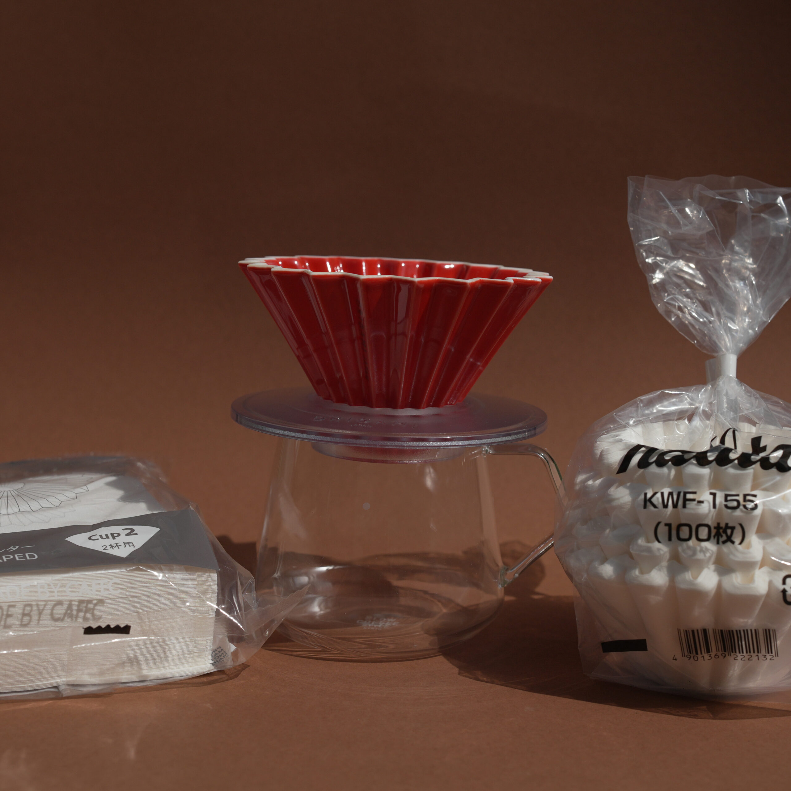 Kaffee Glaskanne mit Origami Filterhalter in rot in der Mitte, rechts und links umgeben von Origami un Kalita Wave Filterpapier