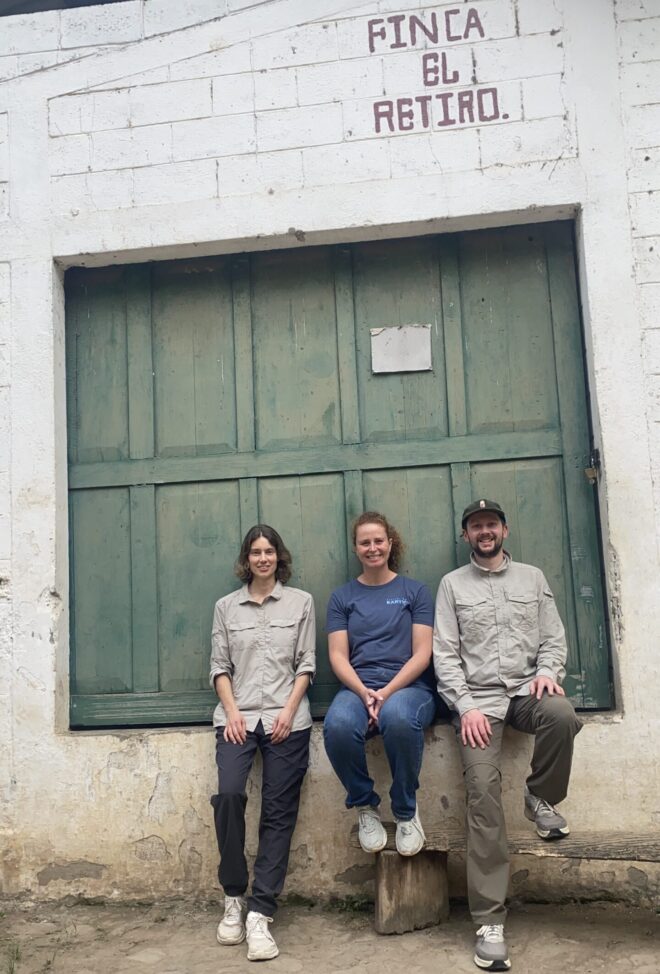 3 Menschen sitzen nebeneinander auf einer Mauer vor einem gruenen Scheunentor